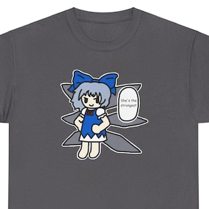 May include: Dark gray t-shirt featuring a cartoon illustration of a character with a blue bow, dress, and hair. The character is inside a stylized shape with the text "She's the strongest" in a speech bubble.