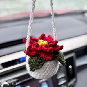May include: A crocheted poinsettia flower in a white pot hangs from the rear view mirror of a car. The flower is red with a yellow centre and green leaves.