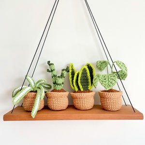 May include: Four crocheted potted plants hanging from a black metal rack. The plants are a snake plant, a ZZ plant, a spider plant, and a monstera. The pots are a light brown color.