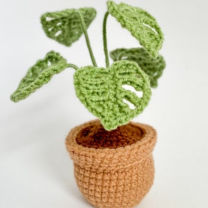 May include: A crocheted green monstera plant in a brown pot. The plant has several leaves and is sitting on a white surface.
