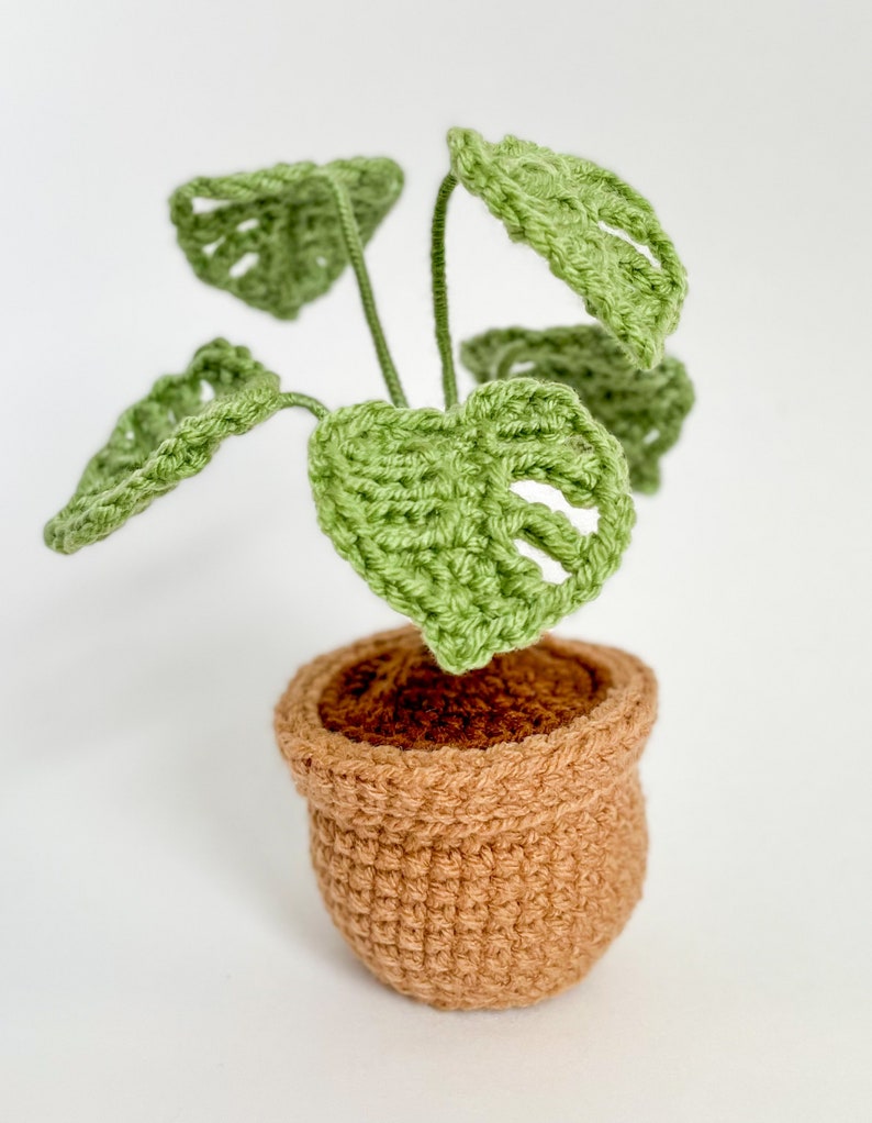 May include: A crocheted green monstera plant in a brown pot. The plant has several leaves and is sitting on a white surface.