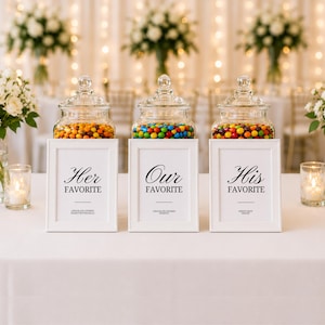 Op de afbeelding: Drie glazen potten gevuld met kleurrijke snoepjes staan op witte ingelijste borden met de tekst "Her Favorite", "Our Favorite" en "His Favorite". De potten staan op een witte tafel, met bloemstukken en kaarsen op de achtergrond.