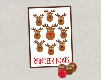 May include: A rectangular card with a brown border featuring nine cartoon reindeer heads on a white background. One reindeer has a bright red nose. The text REINDEER NOSES is printed in red at the bottom. Three brown chocolate candies and one red candy.