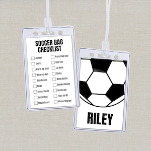 Puede incluir: Dos etiquetas transparentes para bolsas de fútbol colgadas de cordones blancos. Una etiqueta tiene un gráfico de balón de fútbol y el nombre "Riley". La otra etiqueta tiene una lista de verificación de artículos como camisetas, pantalones cortos y espinilleras.
