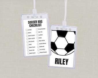 May include: Two clear plastic soccer bag tags hanging by white cords. One tag has a soccer ball graphic and the name "Riley". The other tag has a checklist of items like jerseys, shorts, and shin guards.