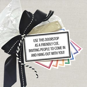 May include: A doorstop gift set wrapped in clear cellophane and tied with a black satin bow with white stitching. A tag reads "USE THIS DOORSTOP AS A FRIENDLY CUE, INVITING PEOPLE TO COME IN AND HANG OUT WITH YOU!" with colorful card accents.
