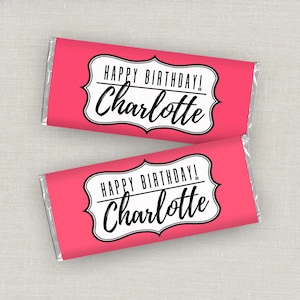 May include: Two pink candy bar wrappers with silver foil ends. Each wrapper has a white and black decorative label that reads "HAPPY BIRTHDAY!" and "Charlotte". The wrappers are on a light gray surface.