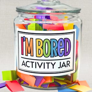 Op de afbeelding: Een doorzichtige glazen pot met deksel, gevuld met kleurrijke papieren strookjes. Een wit rechthoekig etiket op de voorzijde bevat de tekst I'M BORED ACTIVITY JAR in veelkleurige letters, met wat papieren strookjes verspreid rond de pot.
