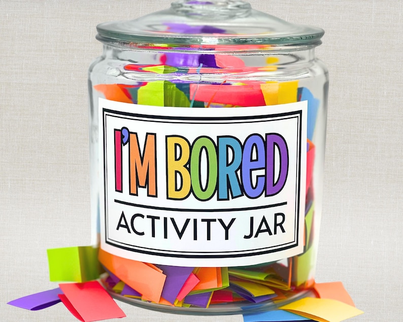Op de afbeelding: Een doorzichtige glazen pot met deksel, gevuld met kleurrijke papieren strookjes. Een wit rechthoekig etiket op de voorzijde bevat de tekst I'M BORED ACTIVITY JAR in veelkleurige letters, met wat papieren strookjes verspreid rond de pot.