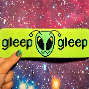 May include: A neon green rectangle with the word "gleep" on each side of a pixelated alien face. The background is a starry galaxy. The image is held by a hand with a matching alien nail design.