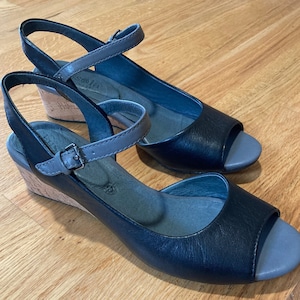 May include: A pair of black leather wedge sandals with gray accents. The sandals have an open toe design, adjustable ankle straps, and cork-style wedge heels. The shoes are displayed on a wooden surface.