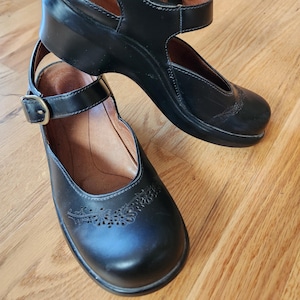 May include: Black leather Mary Jane style shoes with a buckle closure and a decorative floral design on the toe. The shoes have a wedge heel and a strap that goes around the ankle.