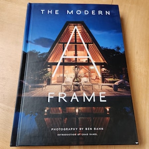 May include: A hardcover book titled "THE MODERN FRAME" with a photograph of a modern A-frame house on the cover. The cover features a large white "A" over the house, with the photographer's name and introduction details below.
