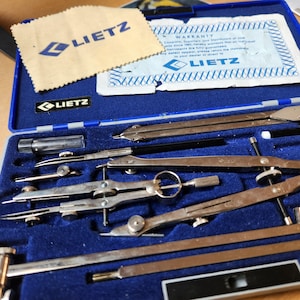 May include: A blue case containing a set of LIETZ drafting tools. The set includes various compasses, dividers, and other precision instruments. A beige cloth with the LIETZ logo and a warranty card are also visible.