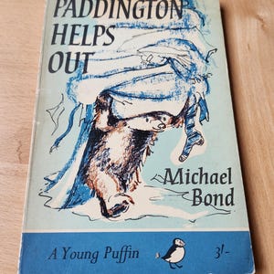 May include: A vintage copy of "Paddington Helps Out" by Michael Bond. The cover shows a blue and brown illustration of Paddington Bear. The book is part of the "A Young Puffin" series, with a price of 3/-.