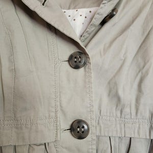 May include: Close-up of a light tan jacket with large, dark buttons. The jacket has a collared neckline and a pleated design at the waist. The interior lining is white with small brown polka dots.