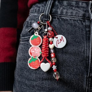 May include: A keychain featuring a silver ring with multiple attachments including a vertical row of two strawberry charms and a flower charm, a red braided cord with a white heart, and a beaded strand with a white disc printed with RM.