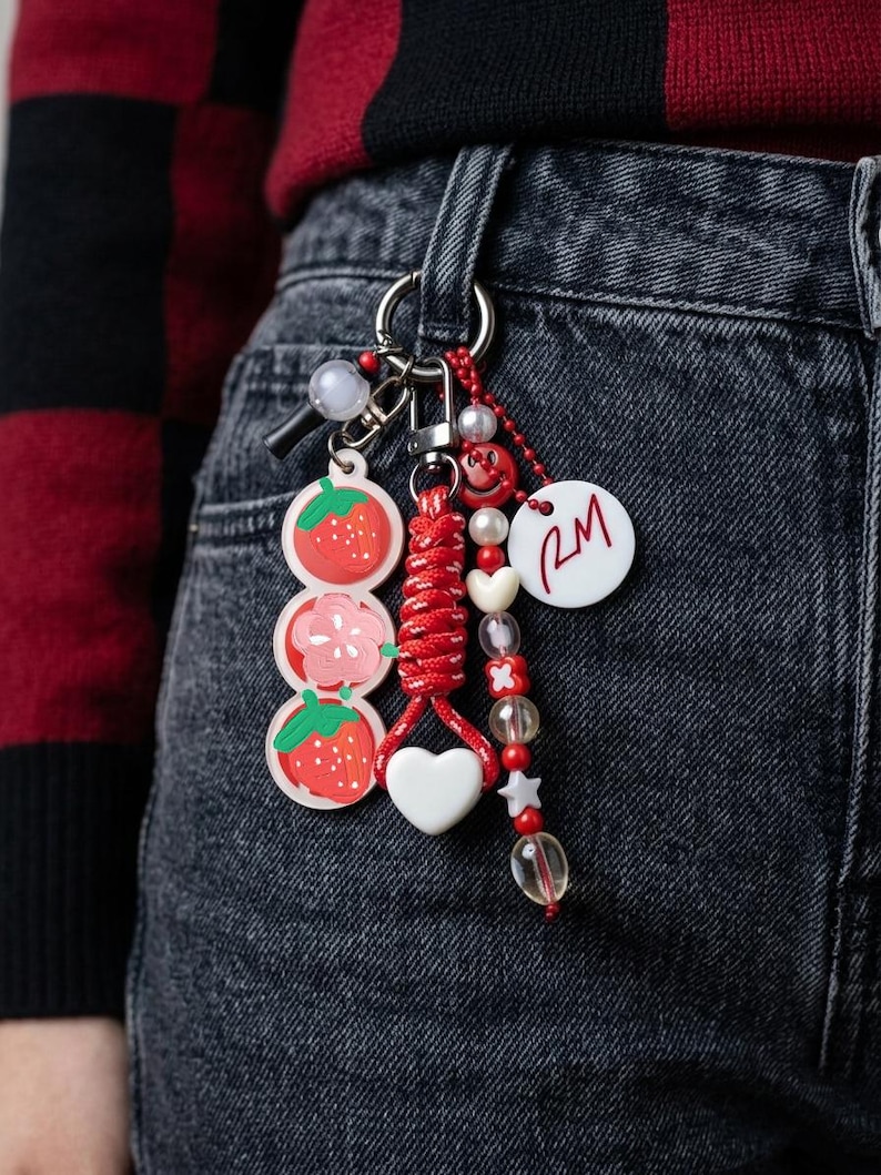 May include: A keychain featuring a silver ring with multiple attachments including a vertical row of two strawberry charms and a flower charm, a red braided cord with a white heart, and a beaded strand with a white disc printed with RM.