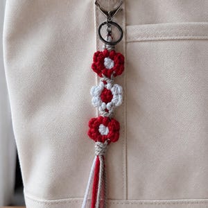 May include: A macrame keyring featuring three crocheted flower motifs in alternating red and white colours, attached to a silver-toned metal lobster clasp and ring. The keyring ends in a multi-coloured tassel made of red, white, and beige yarn strands.