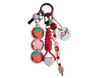 May include: A keychain featuring a vertical charm with two strawberries and a flower, a red braided cord with a white heart, and a beaded strand with a red smiley face, small beads, and a white circular disc with the red letters RM printed on it.