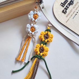 May include: Two macrame keychains with flower designs. One features white daisies with yellow centers, and the other has yellow sunflowers with brown centers. Both have gold-colored clasps and colorful hanging cords. The background includes a wooden tag with text.