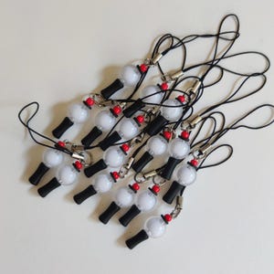 May include: A collection of black phone charms featuring a white spherical bead, a small red accent bead, and a black cylindrical component attached to a black cord loop with a silver-toned metal jump ring and connector.