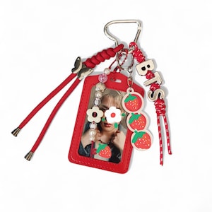 May include: A red rectangular card holder featuring a photo of a person, decorated with a red braided cord, a silver butterfly charm, a beaded strand with white flower charms, and a dangling charm with three red strawberries and the letters BTS.