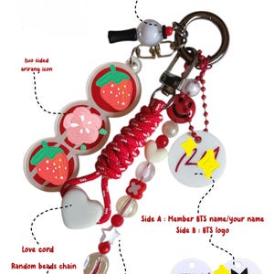 May include: A decorative keychain featuring a red braided cord, a white heart charm, and a random bead chain with red and clear beads. It includes a mini army bomb charm, a two-sided arirang icon with strawberry and flower designs, and a white circular tag.