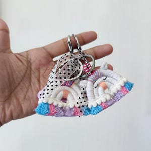 Op de afbeelding: Handgemaakte macramé sleutelhangers met een regenboog en een eenhoorn ontwerp. De sleutelhangers hebben een witte basis met pastel regenboogkleuren, blauwe en paarse franjes en een stippenlint. De sleutelhangers zijn bevestigd aan zilveren sleutelringen.