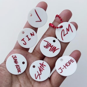 May include: Seven circular white acrylic tags featuring red lettering. The tags display the names V, JIN, JIMIN, RM, JUNG KOOK, SUGA, and J-HOPE. One tag is attached to a red ball chain. Each tag has a small circular hole near the top edge.