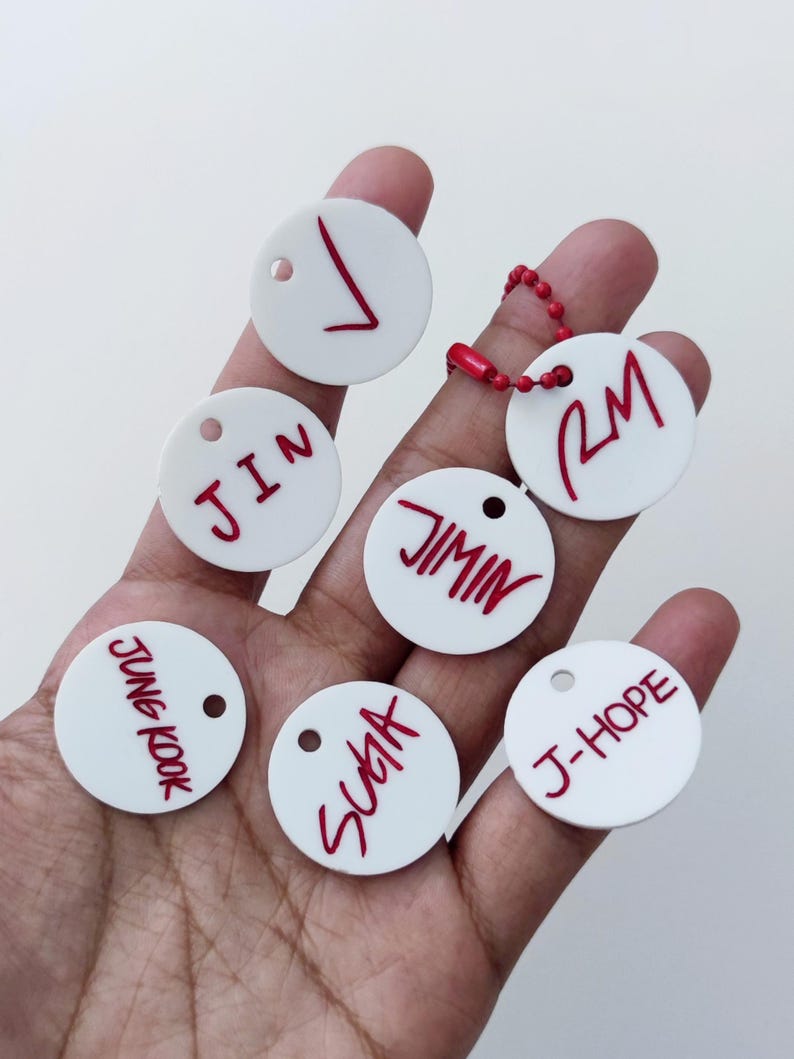 May include: Seven circular white acrylic tags featuring red lettering. The tags display the names V, JIN, JIMIN, RM, JUNG KOOK, SUGA, and J-HOPE. One tag is attached to a red ball chain. Each tag has a small circular hole near the top edge.