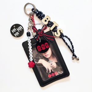 May include: A black rectangular card holder with a clear window containing a photo, attached to a metal keyring. It features a black and white cord, silver letter beads spelling BTS, a white bead strand spelling HOOLIGAN, and red accents.