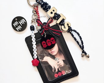 May include: A black rectangular card holder with a clear window containing a photo, attached to a metal keyring. It features a black and white cord, silver letter beads spelling BTS, a white bead strand spelling HOOLIGAN, and red accents.
