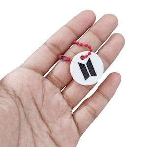 May include: A circular white charm featuring a black BTS logo printed on the front. The charm is attached to a small, bright red ball chain connector, providing a secure loop for attachment to various items or accessories for personal use.