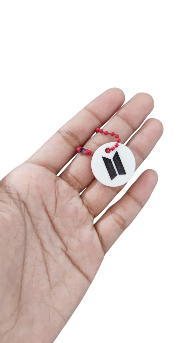 May include: A circular white charm featuring a black BTS logo printed on the front. The charm is attached to a small, bright red ball chain connector, providing a secure loop for attachment to various items or accessories for personal use.