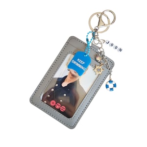 May include: A grey rectangular ID card holder with a clear window containing a photograph. Attached to the metal keyring are charms including a blue tag with the text KEEP SWIMMING, a gold ship wheel, a lifebuoy, a heart, and beads spelling JOON.