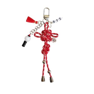 May include: A red and white knotted cord keychain featuring a silver metal lobster clasp, a small red tassel, a white spherical charm with a black base, a silver flower charm, and a string of white beads spelling out the letters B T S.