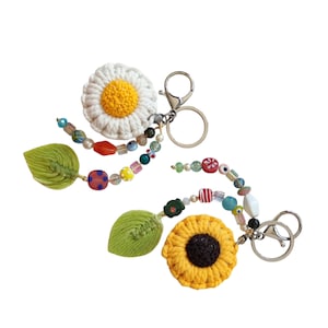 May include: Two handmade keychains featuring crocheted flower designs. One keychain has a white daisy with a yellow center, the other has a yellow sunflower with a brown center. Both have green leaves and colorful beaded accents.