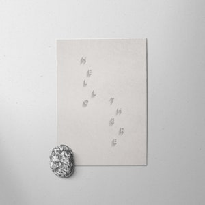 Pode incluir: Um cartão branco com a palavra "silêncio" cortada em uma linha vertical. A palavra está escrita em minúsculas. Uma pequena pedra cinza salpicada fica no canto inferior esquerdo da imagem.