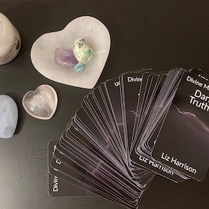 May include: A collection of items on a dark surface, including a deck of black cards with the text "Divine Masculine Dark Truths" and "Liz Harrison". There are heart-shaped crystals and a heart-shaped dish with crystals.