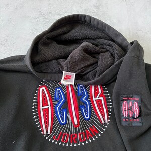 Puede incluir: Sudadera con capucha negra con un dise&ntilde;o bordado en rojo, blanco y azul que presenta el n&uacute;mero "23" y la palabra "JORDAN". La sudadera tambi&eacute;n tiene un peque&ntilde;o parche negro con el texto "AUTHENTIC AIR JORDAN BY NIKE".