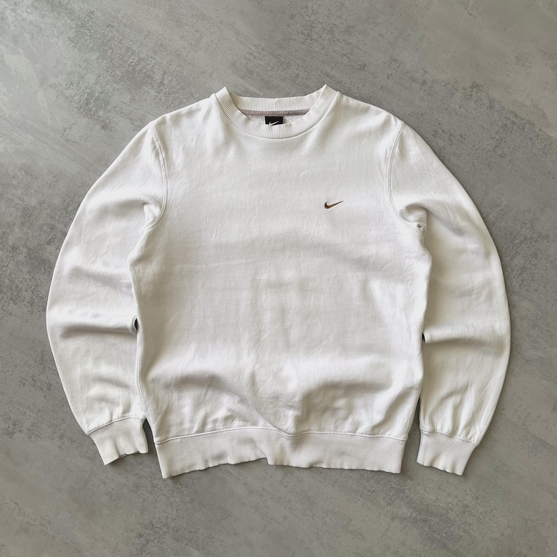 Puede incluir: Sudadera blanca de cuello redondo y manga larga. La sudadera presenta un peque&ntilde;o logotipo marr&oacute;n de Nike en la parte superior izquierda del pecho. Los pu&ntilde;os y el dobladillo son acanalados. El fondo es una superficie gris claro.