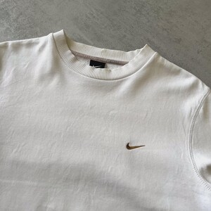 Puede incluir: Sudadera color crema con cuello redondo y un peque&ntilde;o logotipo de Nike marr&oacute;n en el pecho. La sudadera tiene cuello y pu&ntilde;os acanalados, y costuras visibles a lo largo de las costuras. La tela parece ser un material suave y c&oacute;modo.