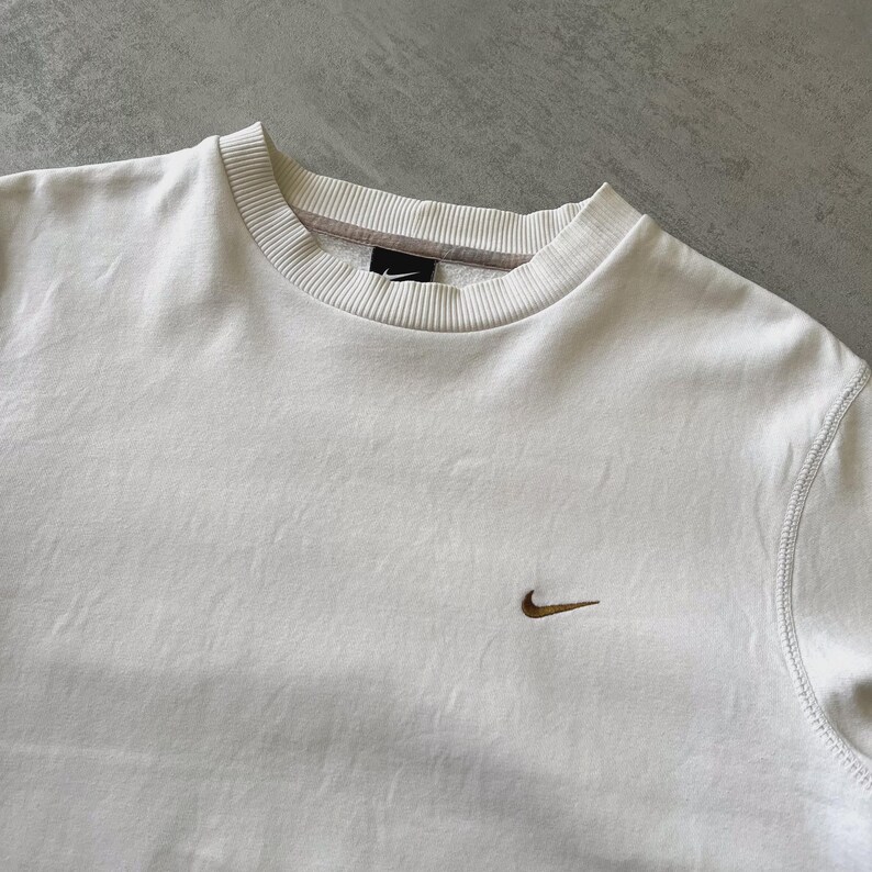 Puede incluir: Sudadera color crema con cuello redondo y un peque&ntilde;o logotipo de Nike marr&oacute;n en el pecho. La sudadera tiene cuello y pu&ntilde;os acanalados, y costuras visibles a lo largo de las costuras. La tela parece ser un material suave y c&oacute;modo.