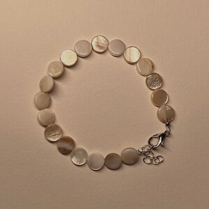 May include: A bracelet featuring round, iridescent, mother-of-pearl beads. The beads are connected by a silver-colored chain and clasp. The bracelet is displayed on a light beige surface.