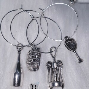 May include: Set of four silver wine glass charms featuring a wine bottle, a wine barrel, a corkscrew, and a wine glass. The charms are attached to silver hoops.