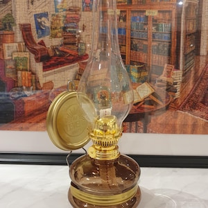 Puede incluir: Una lámpara de aceite de vidrio marrón con una parte superior de metal dorado y una chimenea de vidrio transparente. La lámpara tiene una base de metal dorado y un asa de metal dorado. La lámpara está sobre una superficie blanca. La lámpara tiene una etiqueta que dice "DR. LEVER 1907".
