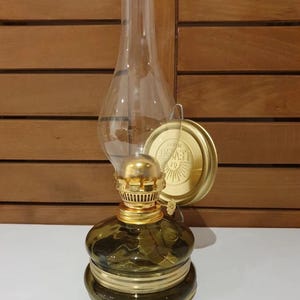 Może przedstawiać: Zielona szklana lampa naftowa z podstawą i kominem ze złotego metalu. Lampa ma złotą metalową pokrywę z napisem "LEVY'S" i wzorem słońca.