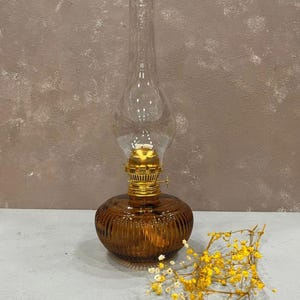 Peut inclure: Une lampe à huile en verre brun avec un cheminée en verre clair et un dessus en métal doré. La lampe est posée sur une surface blanche avec une brindille de fleurs séchées.