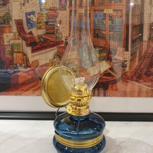 Może przedstawiać: Niebieska szklana lampa naftowa z pozłacanymi zdobieniami i przezroczystym kloszem. Lampa ma mosiężną podstawę i mocowanie ścienne.